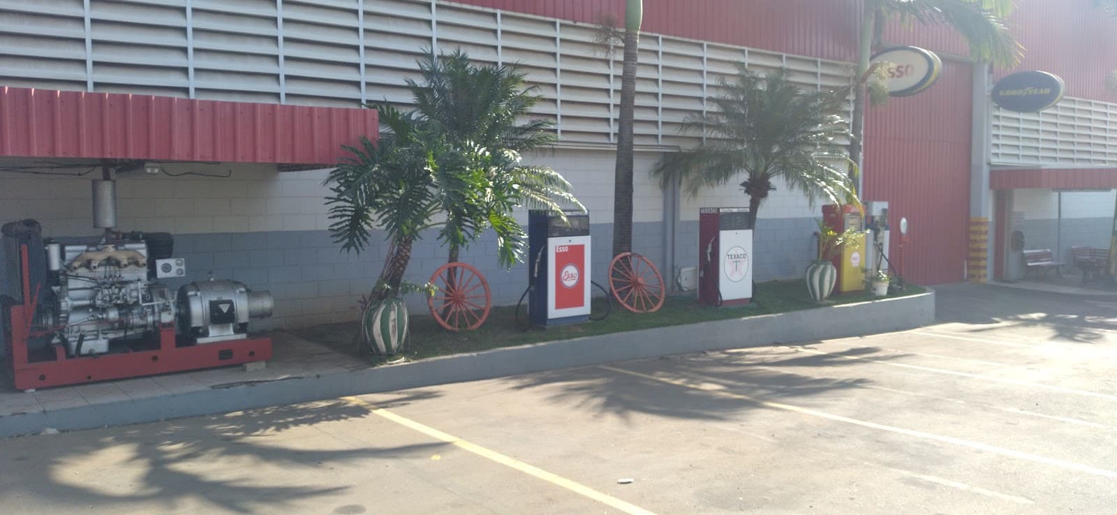 Jardim na frente do galpão com um motor antigo, rodas de carroça e bombas de combustível de época