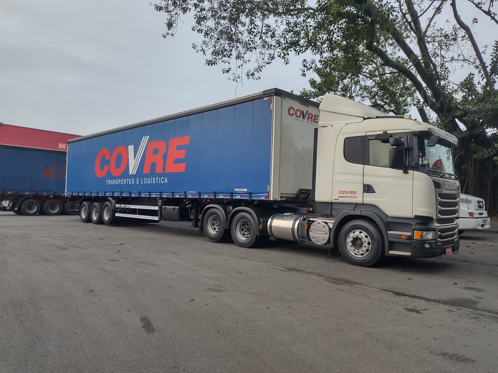 Cavalo Scania branco puxando a carreta de lona azul com o COVRE em vermelho, no pátio da matriz